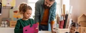A father looks over his young daughters cutting out paper shapes in a crafting room. One daughter sits while the eldest stands.